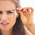 Woman plucking eyebrows while grimmacing.