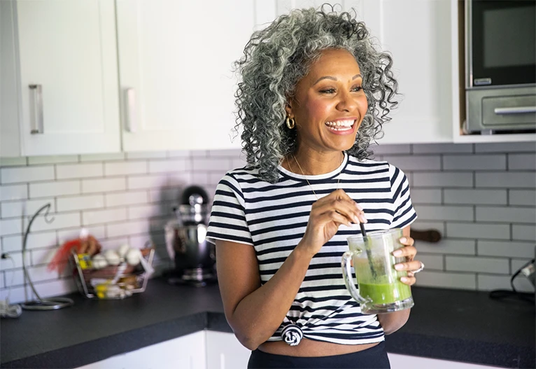 Middle-aged smiling woman drinking a green smoothie in her kitchen.
