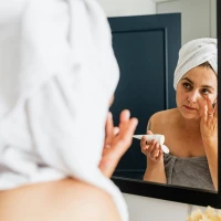 Woman wearing a towel applying face cream in a mirror.