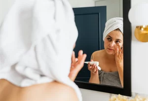 Woman wearing a towel applying face cream in a mirror.